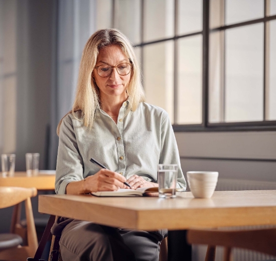 Frau mit Lesebrille sitzt in einem Café und schreibt Tagebuch. 